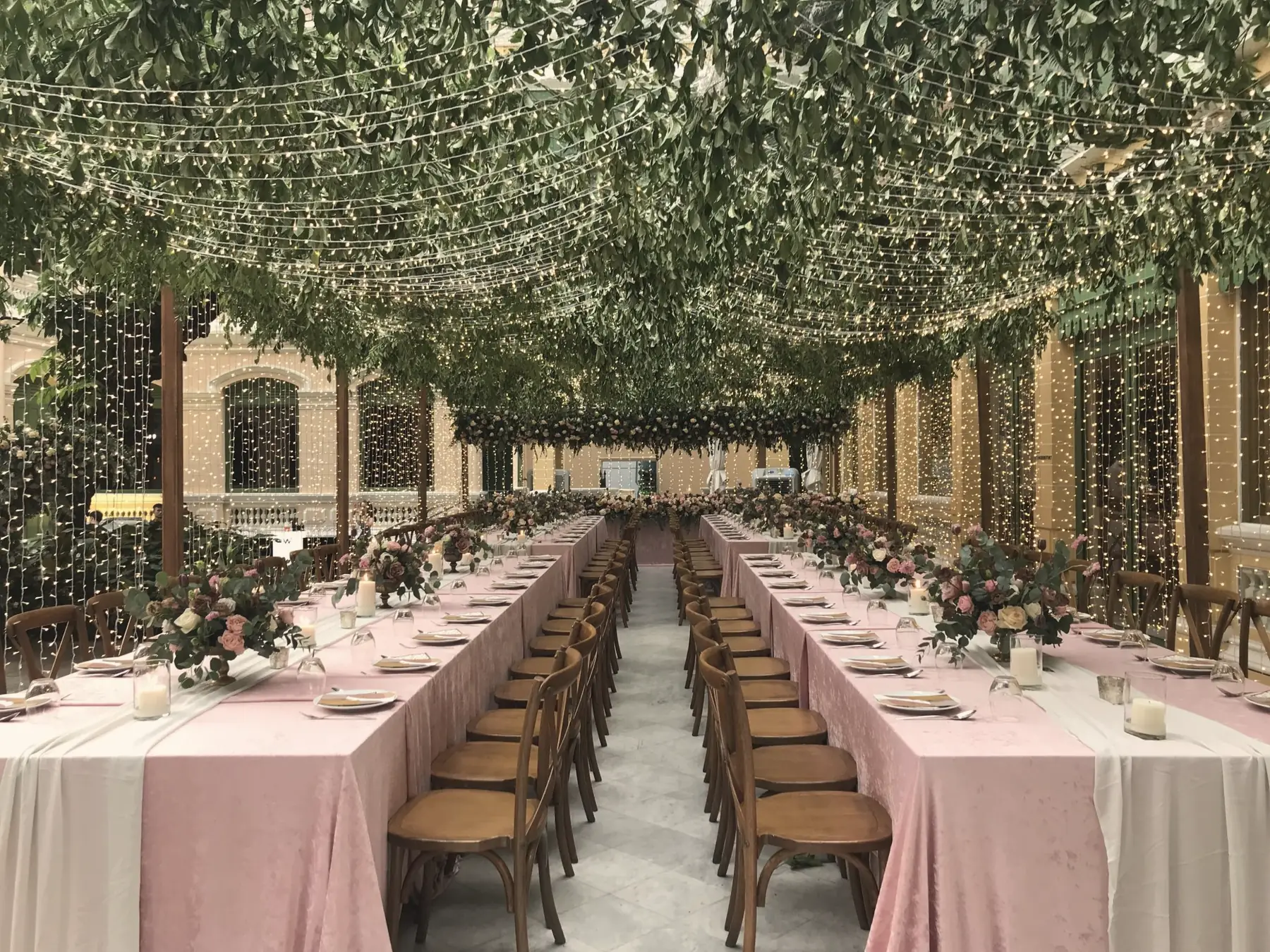 Mesas largas de banquete bajo un techo de hojas y guirnaldas de luces